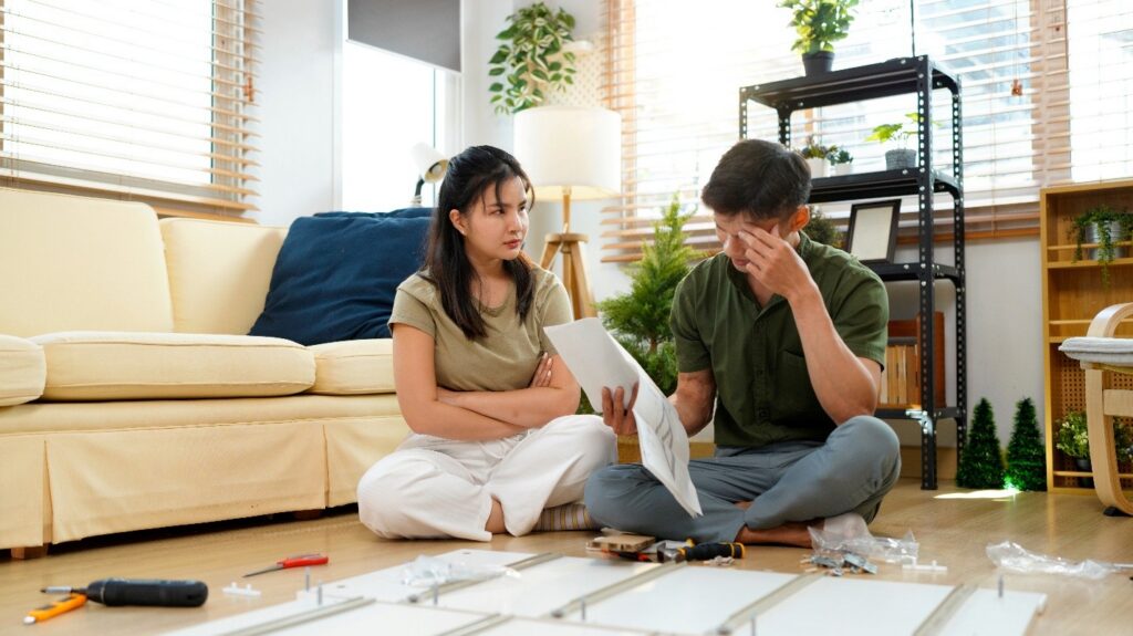 Furniture pieces are arranged on the floor in front of a frustrated man, who is looking in despair at the assembly manual. A woman, sitting next to him, looks on judgmentally.